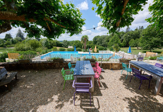 Udendørs spiseområde med farverige stole, borde og udsigt over swimmingpool i grønne omgivelser i Camping Lestaubière.