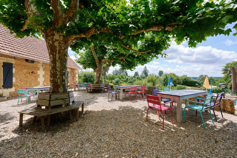 Area esterna con tavoli e sedie colorate sotto alberi ombrosi a Camping Lestaubière in Nouvelle-Aquitaine, Francia.