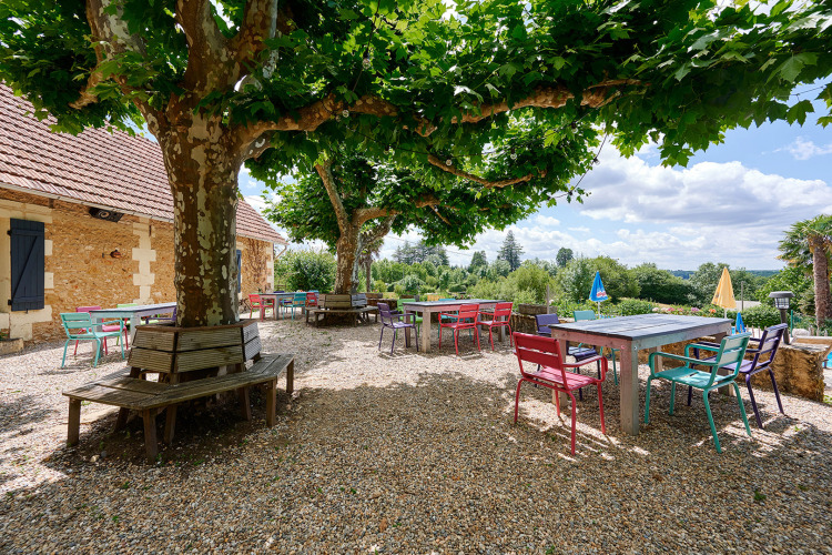 Buitenruimte met kleurrijke tafels en stoelen onder bomen bij Camping Lestaubière in Nouvelle-Aquitaine, Frankrijk.