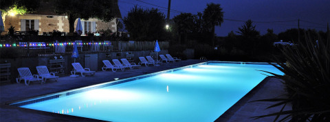 Piscina iluminada en Camping Lestaubière, Nouvelle-Aquitaine, Francia, con tumbonas y terraza por la noche.