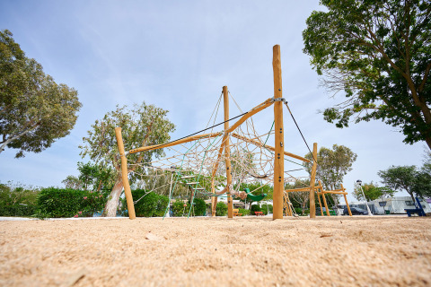 Parque infantil con red de escalada y suelo de arena en Camping Nautic Almata, Cataluña, España, rodeado de árboles.