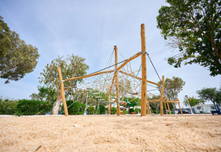 Parque infantil con red de escalada y suelo de arena en Camping Nautic Almata, Cataluña, España, rodeado de árboles.