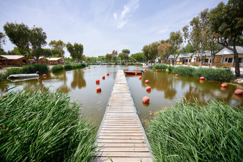 Passerella in legno su uno stagno contornato da bungalow e verde al Camping Nautic Almata in Catalogna, Spagna.