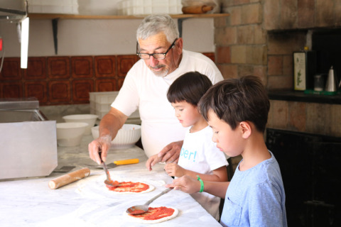 Hombre mayor y dos niños preparando pizza casera juntos en una cocina de Camping Pian Di Boccio en Umbría, Italia.