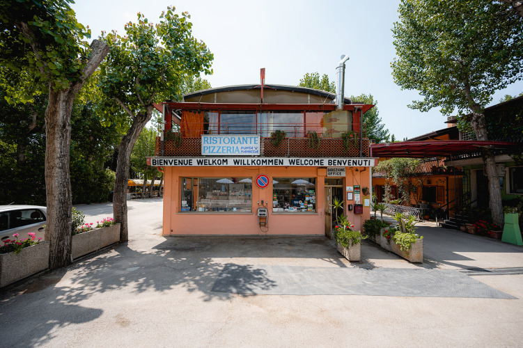 Eingang zu Camping Punta Navaccia mit Restaurant und Büro, umgeben von Bäumen in Umbrien, Italien.