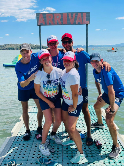 Six amis souriants posent sous une pancarte 'ARRIVAL' au bord du lac du Camping Punta Navaccia, en Ombrie.