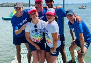 Sei persone sorridenti sotto il cartello ARRIVAL al lago nel Camping Punta Navaccia, Umbria, Italia.