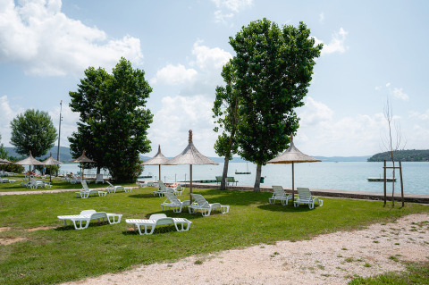 Area campeggio verde sul lago con lettini e ombrelloni di paglia a Camping Punta Navaccia, Umbria, Italia.