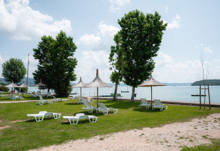 Area campeggio verde sul lago con lettini e ombrelloni di paglia a Camping Punta Navaccia, Umbria, Italia.