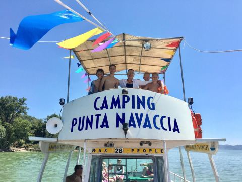 Gruppo di persone su una barca con il cartello Camping Punta Navaccia in una giornata di sole in Umbria, Italia.
