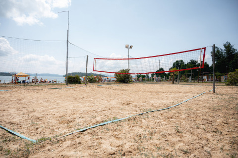 A sandy volleyball court at Camping Punta Navaccia in Umbria, Italy, overlooking the lake with people relaxing nearby.