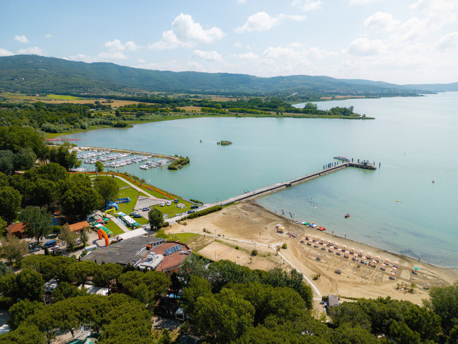 Luftaufnahme vom Camping Punta Navaccia in Umbrien, Italien, mit See, Strand, Steg und Marina bei Sonnenschein.