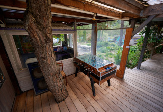 Espace détente extérieur couvert avec baby-foot et arbre traversant la terrasse au Camping River, France.