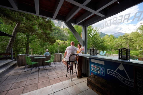 Persone si rilassano con drink su una terrazza tra il verde a Camping River, Provenza-Alpi-Costa Azzurra, Francia.