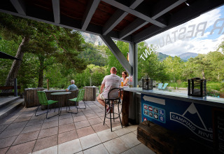 Persone si rilassano con drink su una terrazza tra il verde a Camping River, Provenza-Alpi-Costa Azzurra, Francia.