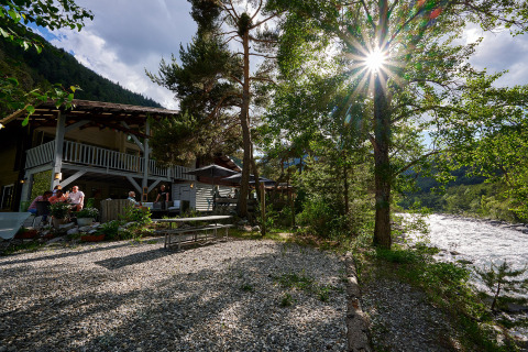Vista exterior en Camping River en Provence-Alpes-Côte d’Azur, con río, sol, árboles y huéspedes en la terraza.