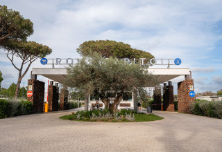 De ingang van Camping Roma Capitol vakantiepark in Lazio, Italië, met bomen en stenen zuilen aan weerszijden.