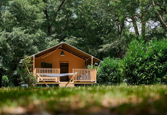 Tienda safari Villatent Nomad en Camping Lestaubière, Francia, rodeada de árboles verdes y una hamaca al frente.