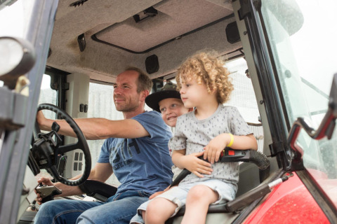 Un uomo e due bambini seduti insieme su un trattore a FarmCamps Op Flakkee, parco vacanze in Olanda Meridionale, Paesi Bassi.