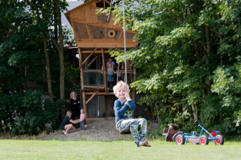 Junge schaukelt vor einem Baumhaus, mehrere Personen im Hintergrund, FarmCamps Op Flakkee, Niederlande.