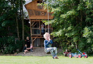Junge schaukelt vor einem Baumhaus, mehrere Personen im Hintergrund, FarmCamps Op Flakkee, Niederlande.