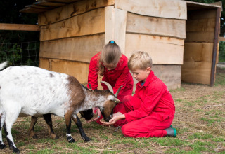 Twee kinderen in rode overall voederen een geit bij een houten hut op FarmCamps Op Flakkee, Zuid-Holland.