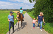 To kvinder og to børn går og rider på en hest på en landevej omgivet af marker, ved FarmCamps Soodehof Eins.