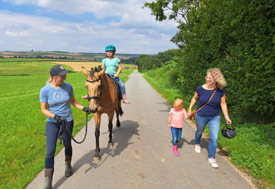 To kvinder og to børn går og rider på en hest på en landevej omgivet af marker, ved FarmCamps Soodehof Eins.
