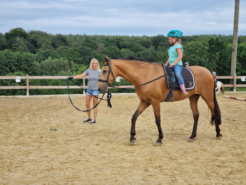 Una niña con casco monta un caballo, guiado por una mujer en una pista en FarmCamps Soodehof Eins, Hesse.