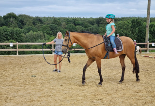A girl wearing a helmet rides a horse led by a woman on a riding arena at FarmCamps Soodehof Eins in Hesse.