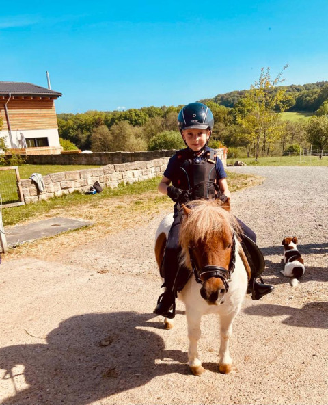 A child wearing a helmet rides a pony at FarmCamps Soodehof Eins in Hesse, Germany, on a sunny day.
