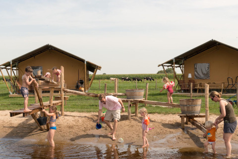 Kinder und Erwachsene spielen am Wasserspielplatz vor Zelthütten im FarmCamps Stolkse Weide, Niederlande.