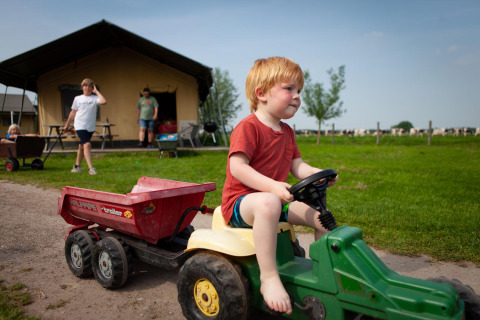 Un niño pequeño conduce un tractor de juguete frente a una cabaña en FarmCamps Stolkse Weide, Holanda Meridional.
