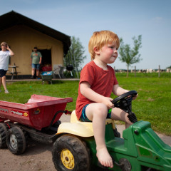 Een jong kind rijdt op een speelgoedtractor bij een huisje op FarmCamps Stolkse Weide in Zuid-Holland.
