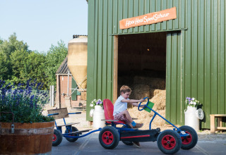 Un garçon conduit un kart devant une grange avec le panneau 'Hooi Hooi Speel Schuur' au FarmCamps Stolkse Weide.