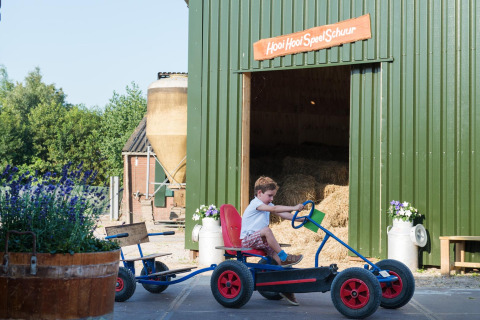 A boy rides a go-kart in front of a barn with the sign 'Hooi Hooi Speel Schuur' at FarmCamps Stolkse Weide.