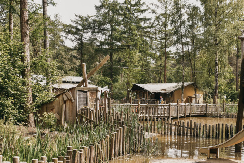 Wooden cabins and a boardwalk beside a pond surrounded by greenery at Camping het Horstmannsbos, Drenthe.