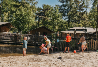 Des enfants jouent près du lac à Camping het Horstmannsbos, Drenthe, Pays-Bas, entourés d’arbres et cabanes.