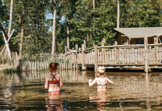 Two children wading in a natural pond at Camping het Horstmannsbos, with wooden cabins and trees behind.