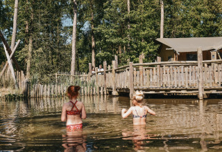 Two children wading in a natural pond at Camping het Horstmannsbos, with wooden cabins and trees behind.