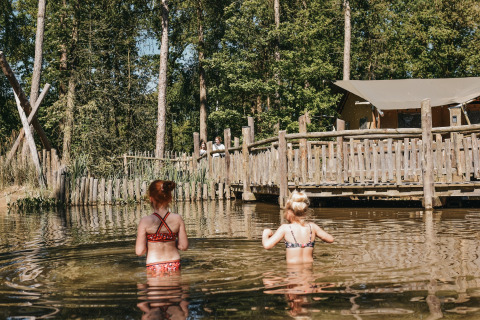 Two children wading in a natural pond at Camping het Horstmannsbos, with wooden cabins and trees behind.