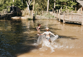 Due bambini giocano nell’acqua al Camping het Horstmannsbos, circondati da alberi e recinzioni in legno.