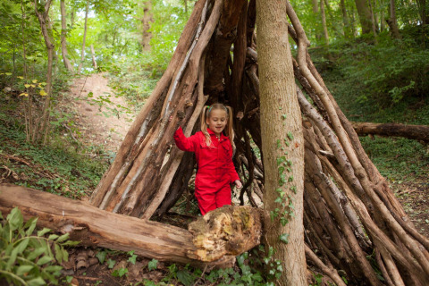 Child in a red playsuit exploring a stick shelter in the woods at FarmCamps De Verloren Kost, Limburg, Netherlands.