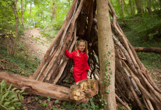 Barn i rød kedeldragt leger i skovhytte lavet af grene i naturen ved FarmCamps De Verloren Kost, Limburg.