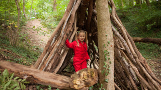 Niña con mono rojo explora un refugio hecho de ramas en el bosque de FarmCamps De Verloren Kost, Limburg.