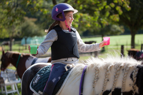 Girl in a helmet and vest riding a horse, holding colorful cups at FarmCamps De Verloren Kost, Limburg, Netherlands.