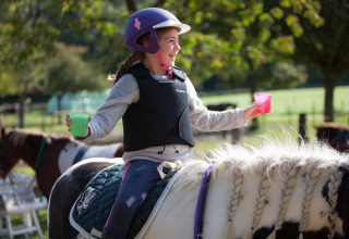 Mädchen mit Helm und Schutzweste reitet auf Pferd, hält zwei bunte Becher auf FarmCamps De Verloren Kost, Limburg.