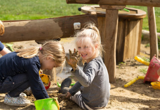 Zwei Kinder spielen lachend im Sand mit Eimern und Spielzeug bei FarmCamps De Verloren Kost in Limburg, Niederlande.
