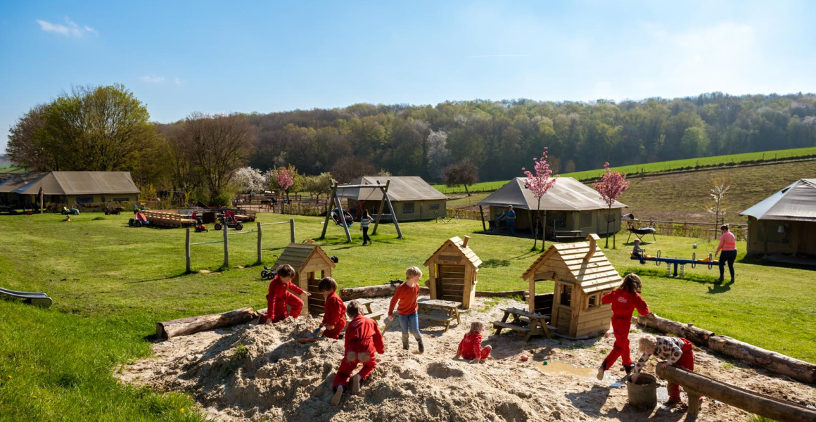 Kinderen spelen in een grote zandbak op FarmCamps De Verloren Kost met grasvelden en tenten op de achtergrond.