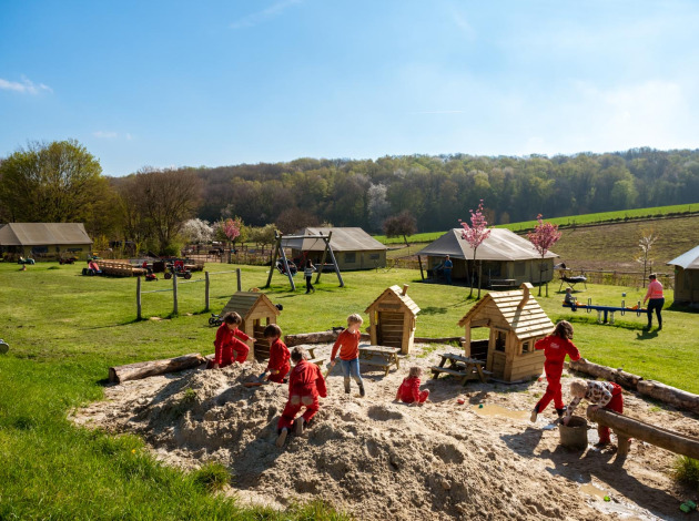 Kinderen spelen in een grote zandbak op FarmCamps De Verloren Kost met grasvelden en tenten op de achtergrond.
